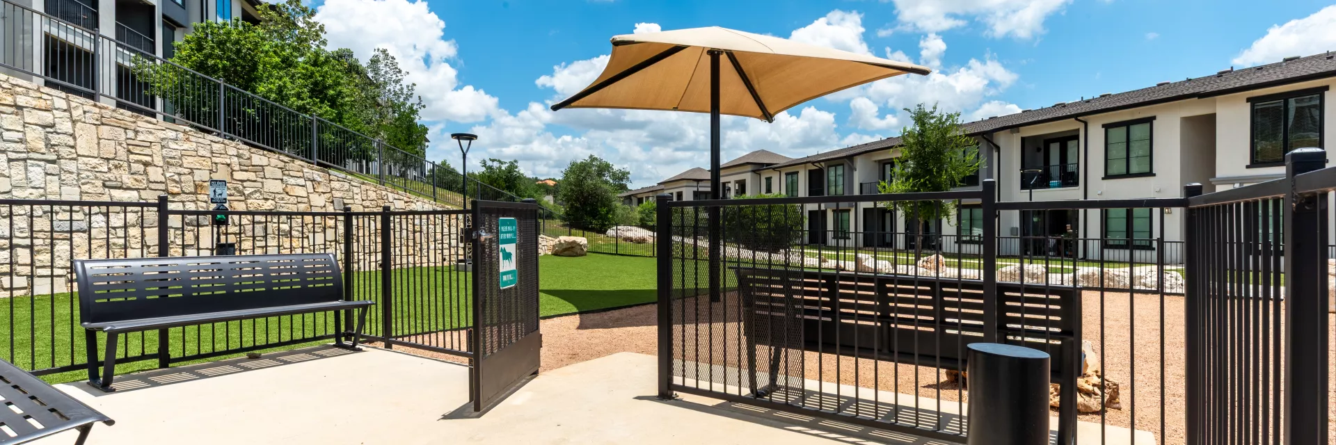 Sunny outdoor dog park entrance with concrete path, black fence, benches, grassy area, shade umbrella, and modern apartments.