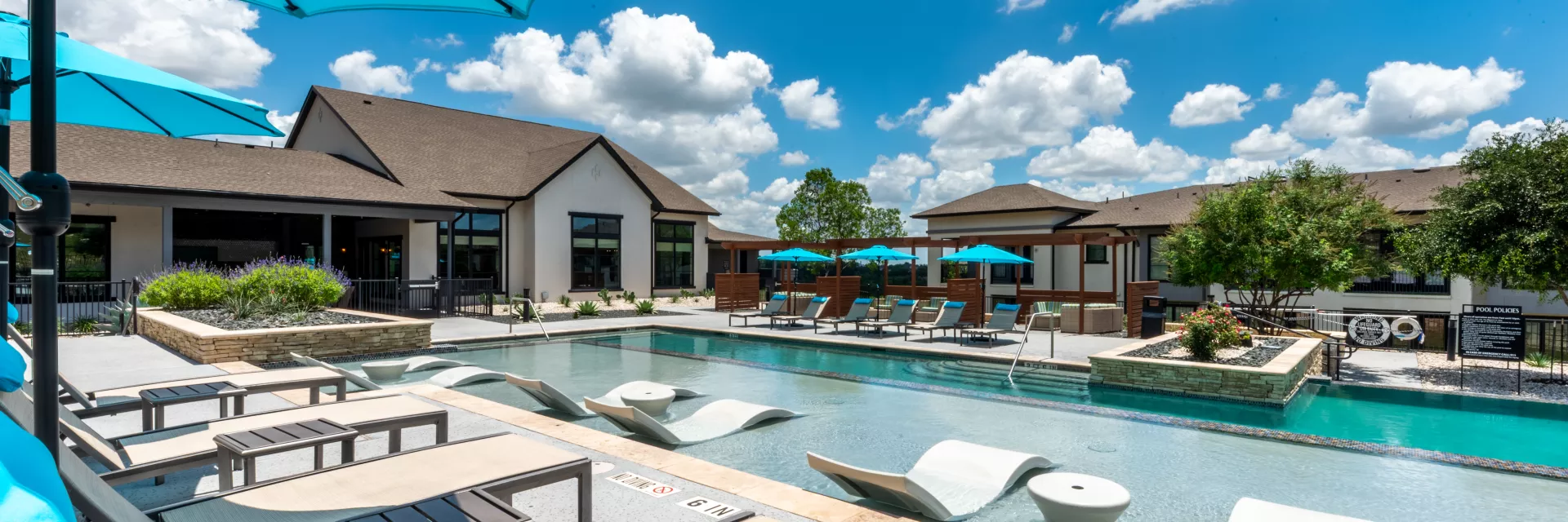 Sunny pool area with turquoise umbrellas, in-water and poolside lounge chairs, and a modern clubhouse building.