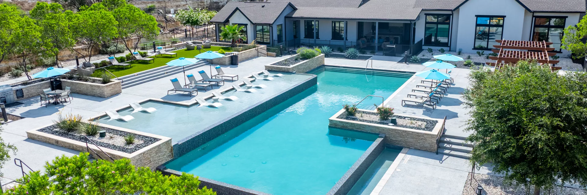 Aerial photograph: Modern clubhouse overlooking a multi-section swimming pool, lounge chairs, blue umbrellas, and lush green hills.
