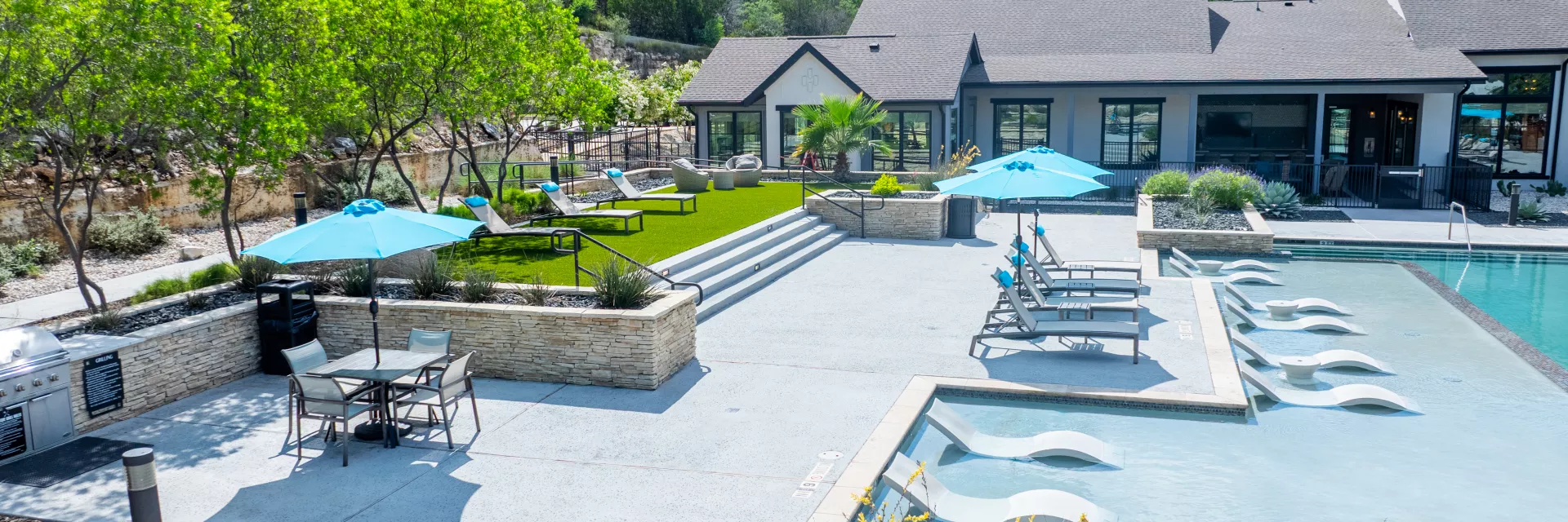 Sunny modern pool area with submerged loungers, blue umbrellas, lush landscaping, and a contemporary clubhouse building.