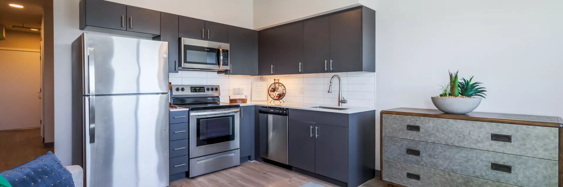 Modern studio apartment with dark gray kitchen cabinets, stainless steel appliances, and a living area featuring a blue rug.