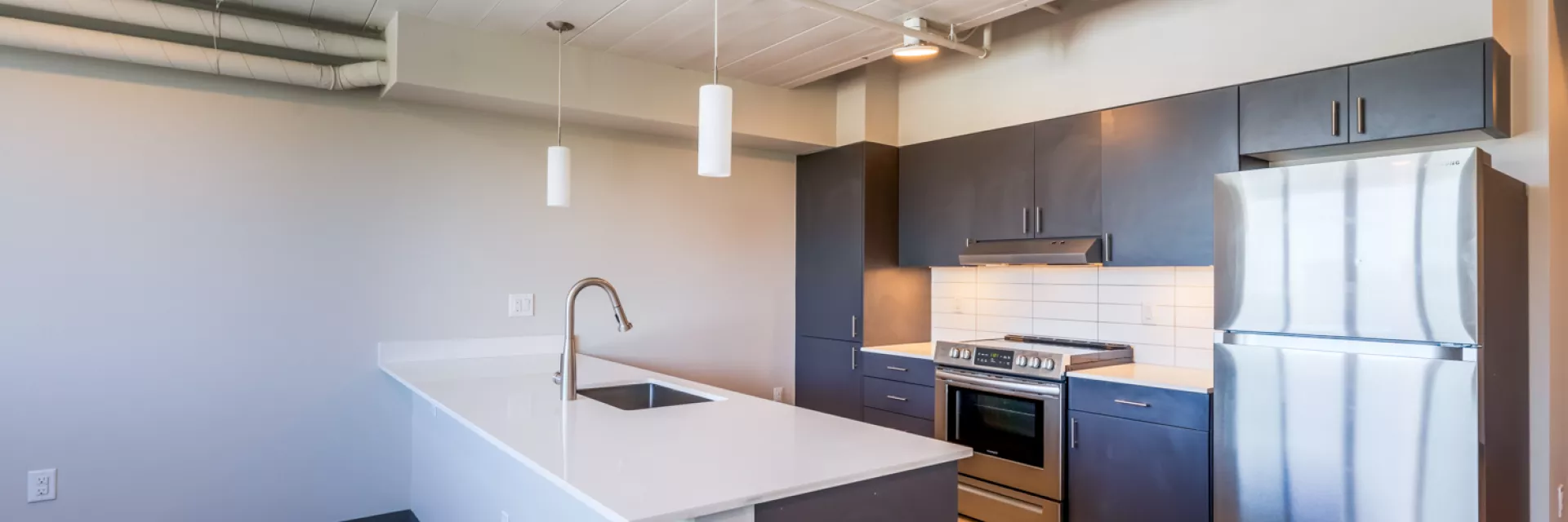 Empty modern kitchen with a white island, sink, dark cabinets, stainless steel appliances, and light wood floor.