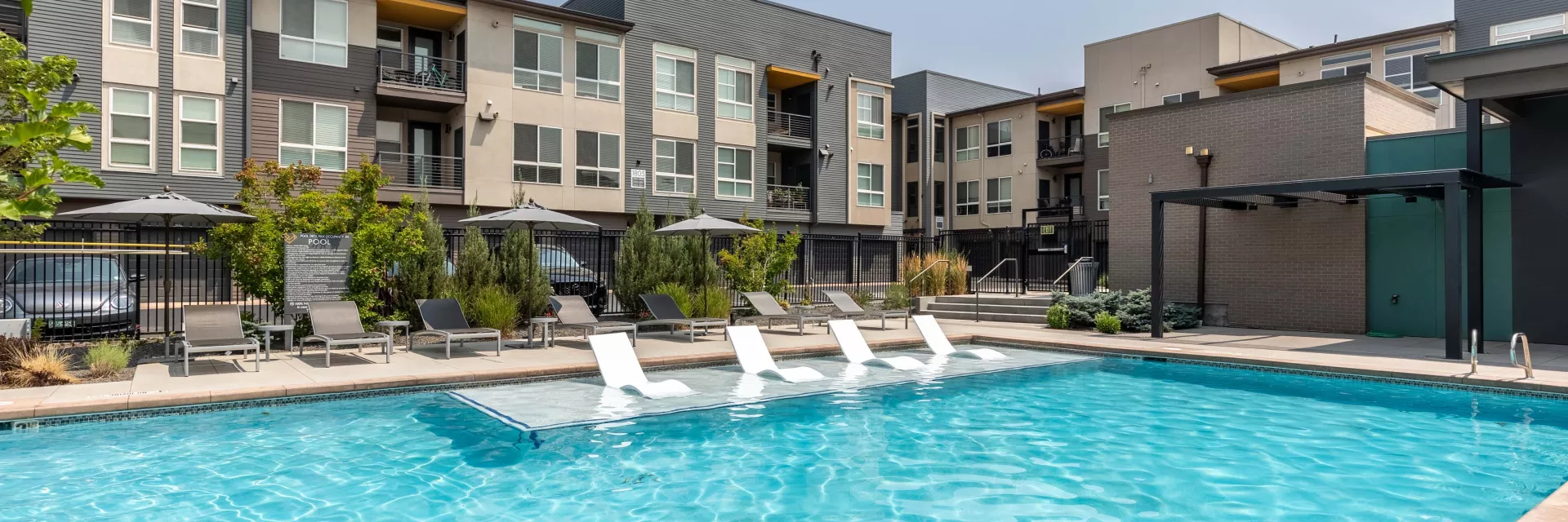 Sunny outdoor swimming pool with in-pool loungers, surrounded by modern apartment buildings with balconies.