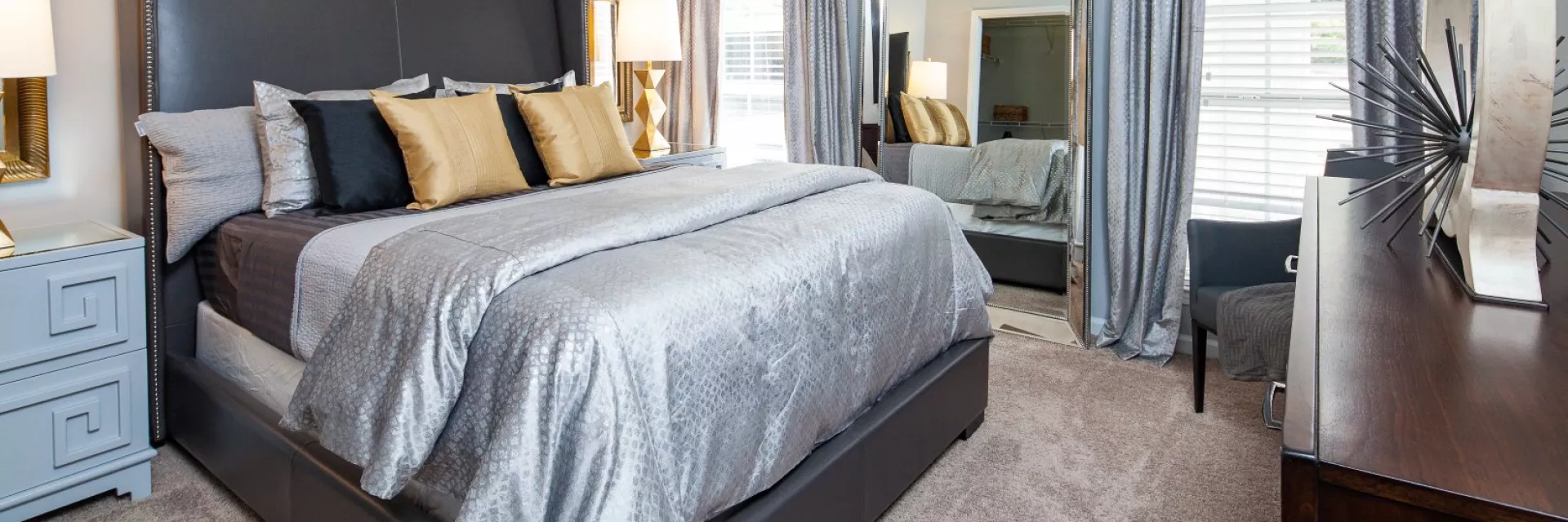 Spacious bedroom with a large bed, grey comforter, gold and black pillows, nightstand, dresser, and full-length mirror.