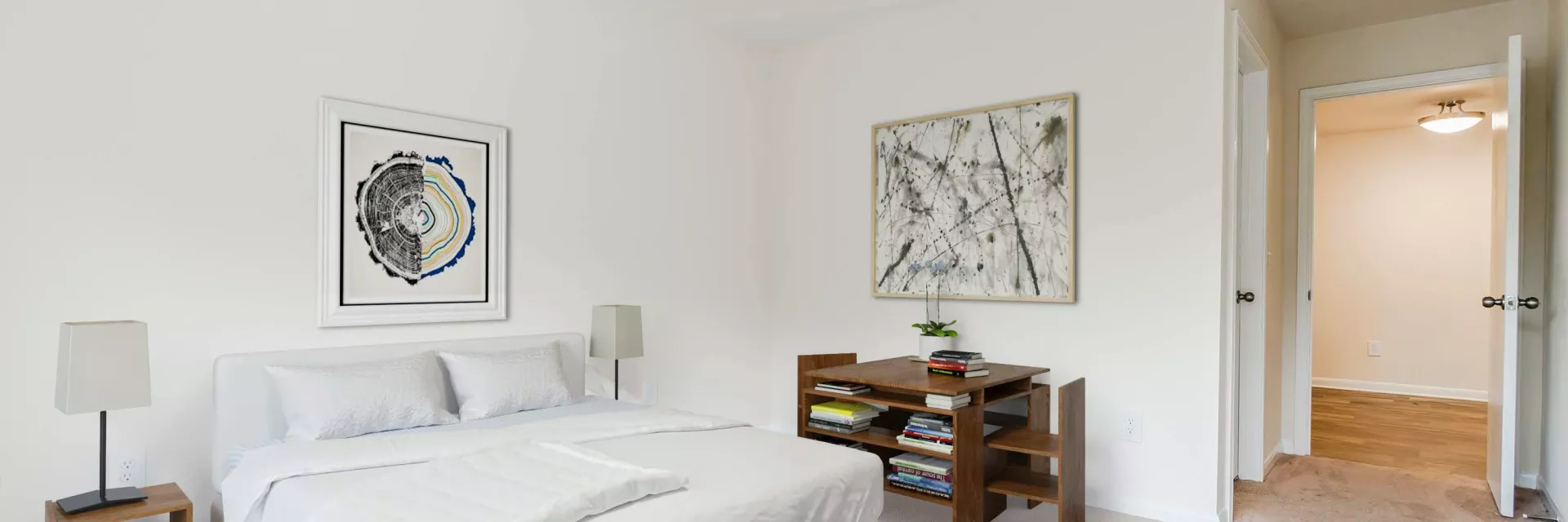 Bright bedroom with a white bed, abstract wall art, wooden desk, and a colorful round rug on light pink carpet.