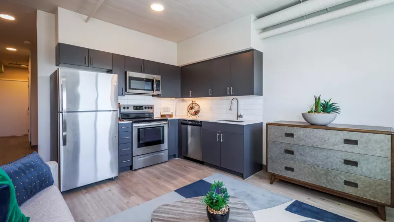 Modern studio apartment with dark gray kitchen cabinets, stainless steel appliances, and a living area featuring a blue rug.