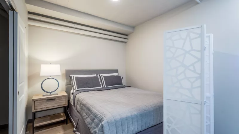 Modern bedroom with a gray bed, light wood nightstand, blue lamp, and a white geometric room divider.