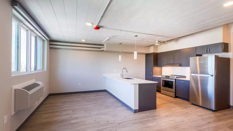 Empty modern apartment with an open kitchen, light wood floors, stainless steel appliances, and a large window.