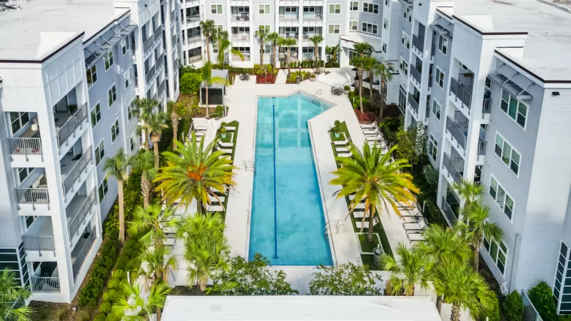 Aerial view of a modern apartment complex with a long blue swimming pool surrounded by palm trees and light-colored buildings.