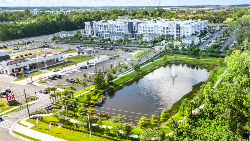 Aerial view of a modern apartment complex, commercial shops, and a lake with a fountain under a blue sky.