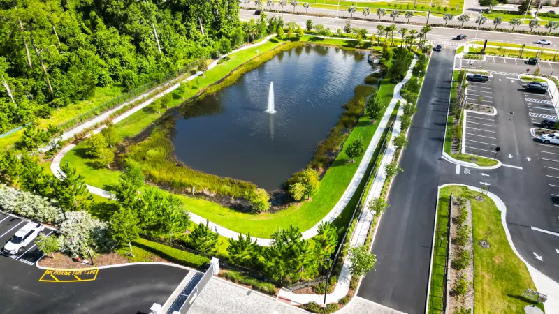 Aerial view of a landscaped park with a pond and fountain, surrounded by green paths, roads, and parking lots.