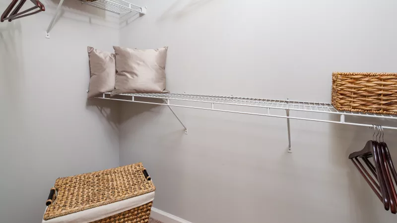 Empty walk-in closet with white wire shelves, wicker baskets, wooden hangers, and two beige pillows.