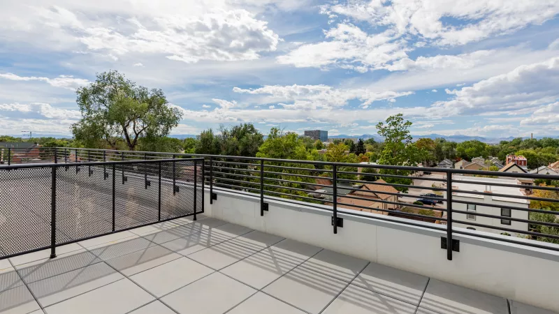 Rooftop deck with light gray tiles and black railing overlooking a city, trees, and distant mountains under a cloudy sky.
