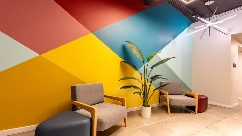 Vibrant lobby with a geometric accent wall in red, yellow, and blue. Two gray chairs, blue ottoman, and a tall plant.
