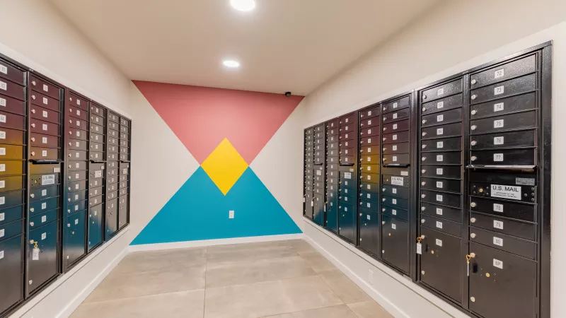 Narrow hallway with dark mailboxes lining walls. Far wall features a geometric mural of pink, blue, and yellow shapes.