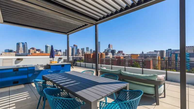 Rooftop patio with a gray table, teal chairs, and a sofa under a pergola, offering a city skyline view.