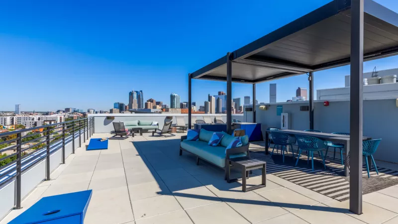 Rooftop patio with modern lounge and dining furniture under a pergola, overlooking a city skyline on a sunny day.