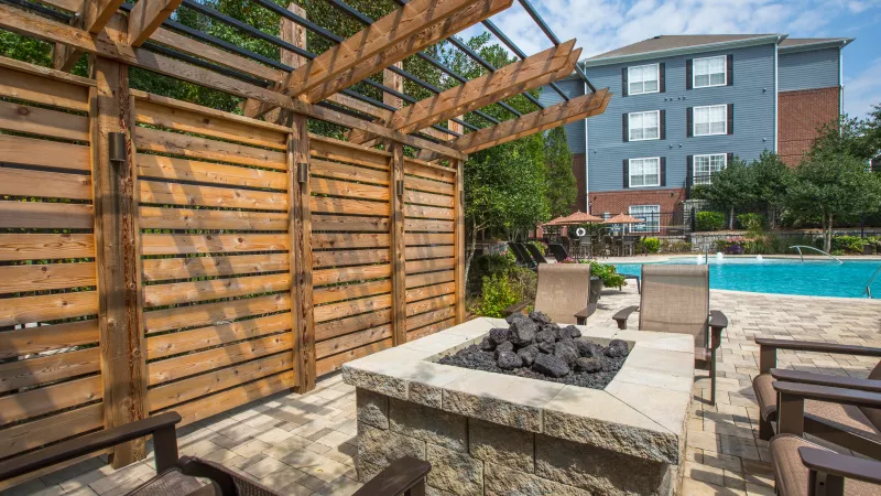 Outdoor patio with stone fire pit, wood pergola, and lounge chairs next to a blue swimming pool and apartment building.