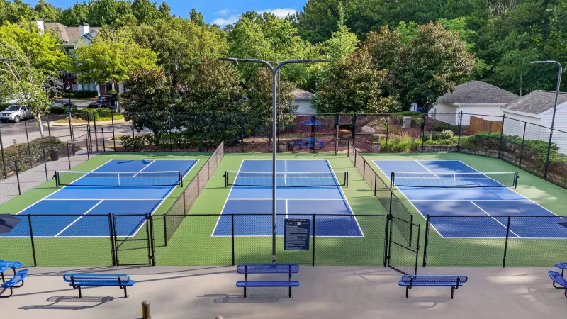 Aerial view of three vibrant blue pickleball courts, fenced, with green turf, benches, and distant houses under a clear sky.