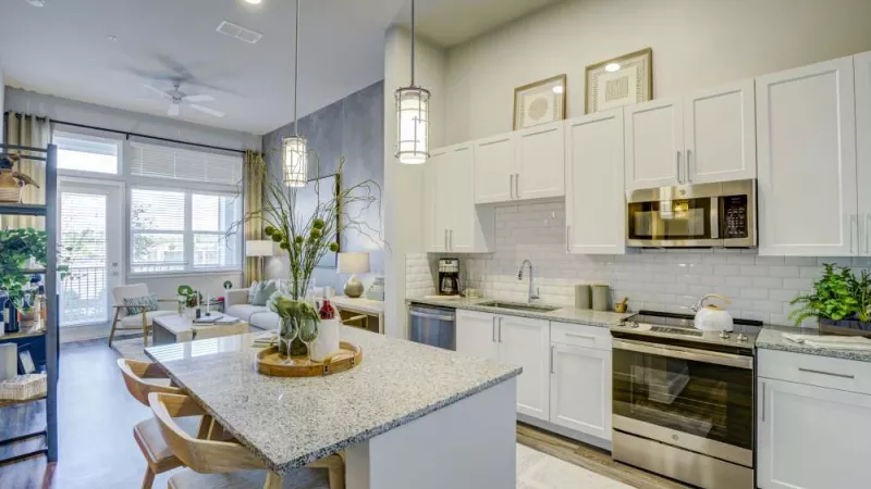 Modern open-concept apartment with a white kitchen, granite island, and a brightly lit living room beyond.
