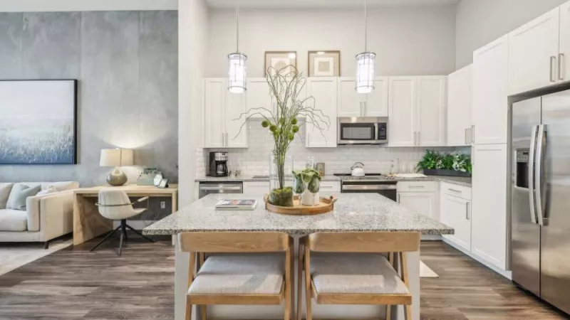 Modern kitchen with white cabinets, granite island, stainless steel appliances, open to a light gray living area.