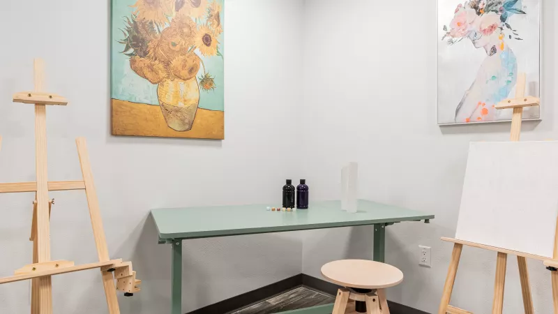Small art studio with two easels, a green desk, stool, Van Gogh's Sunflowers, and a colorful bird painting on walls.