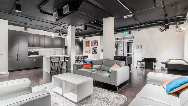Modern common area with a dark kitchenette, grey and white sofas on a rug, and a pool table. Dark, industrial ceiling.