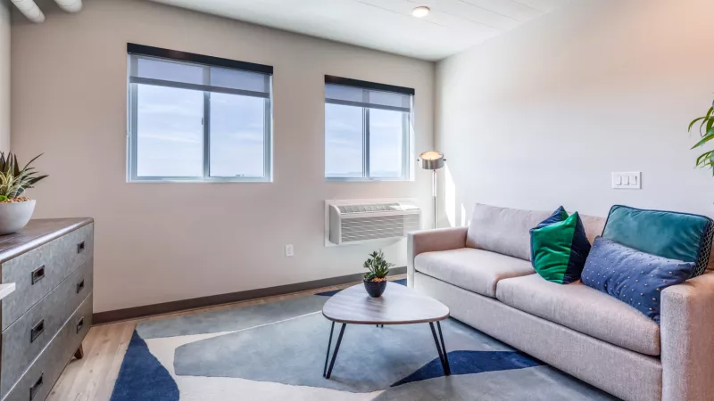 Modern living room with a light sofa, blue geometric rug, coffee table, dresser, and two windows with bright sky views.