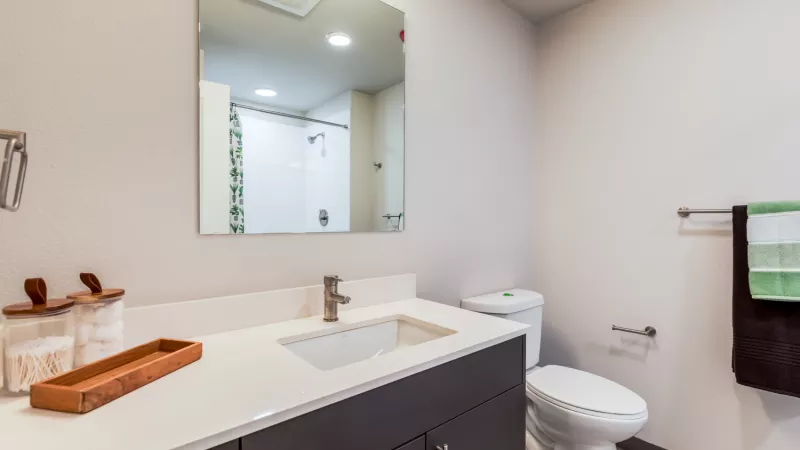 Contemporary bathroom with gray vanity, white countertop, sink, large mirror, white toilet, and hanging green and brown towels.