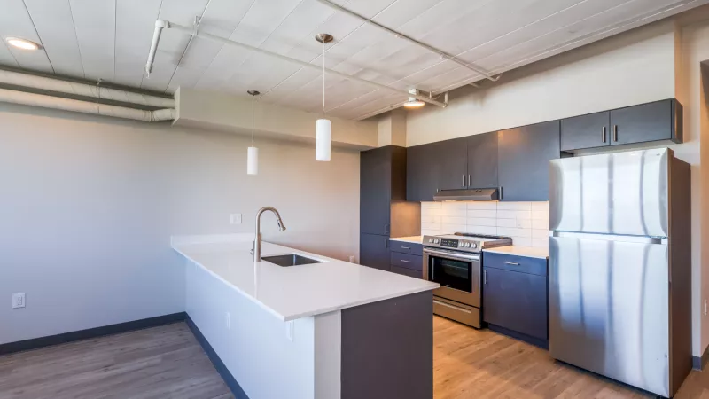 Empty modern kitchen with a white island, sink, dark cabinets, stainless steel appliances, and light wood floor.