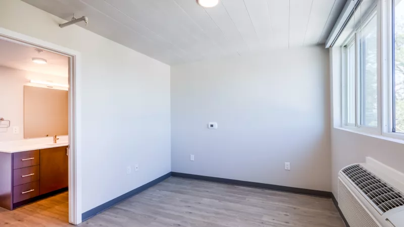 Empty room with a wood-look floor, large window, heating unit, and an open doorway to a bathroom vanity.