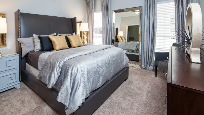 Luxurious bedroom with a large bed, dark headboard, silver bedding, gold and black pillows, and floor mirror.