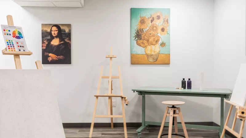 Art studio with wooden easels, blank canvases, a green table holding supplies, and wall prints of Mona Lisa and Van Gogh's Sunflowers.