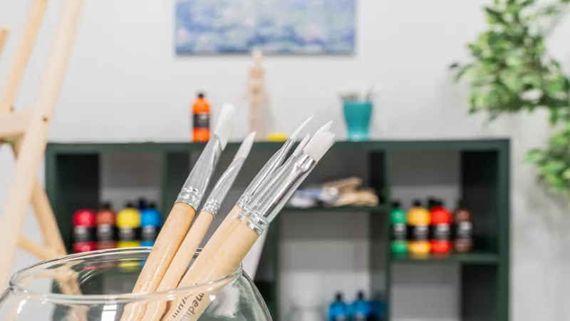 Paintbrushes in a glass, with colorful paint bottles on shelves and a blurred landscape painting in an art studio.