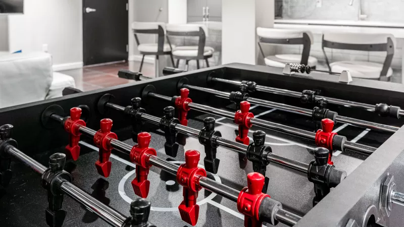 Modern foosball table with red and black players, set in a game room with a bar and stools.