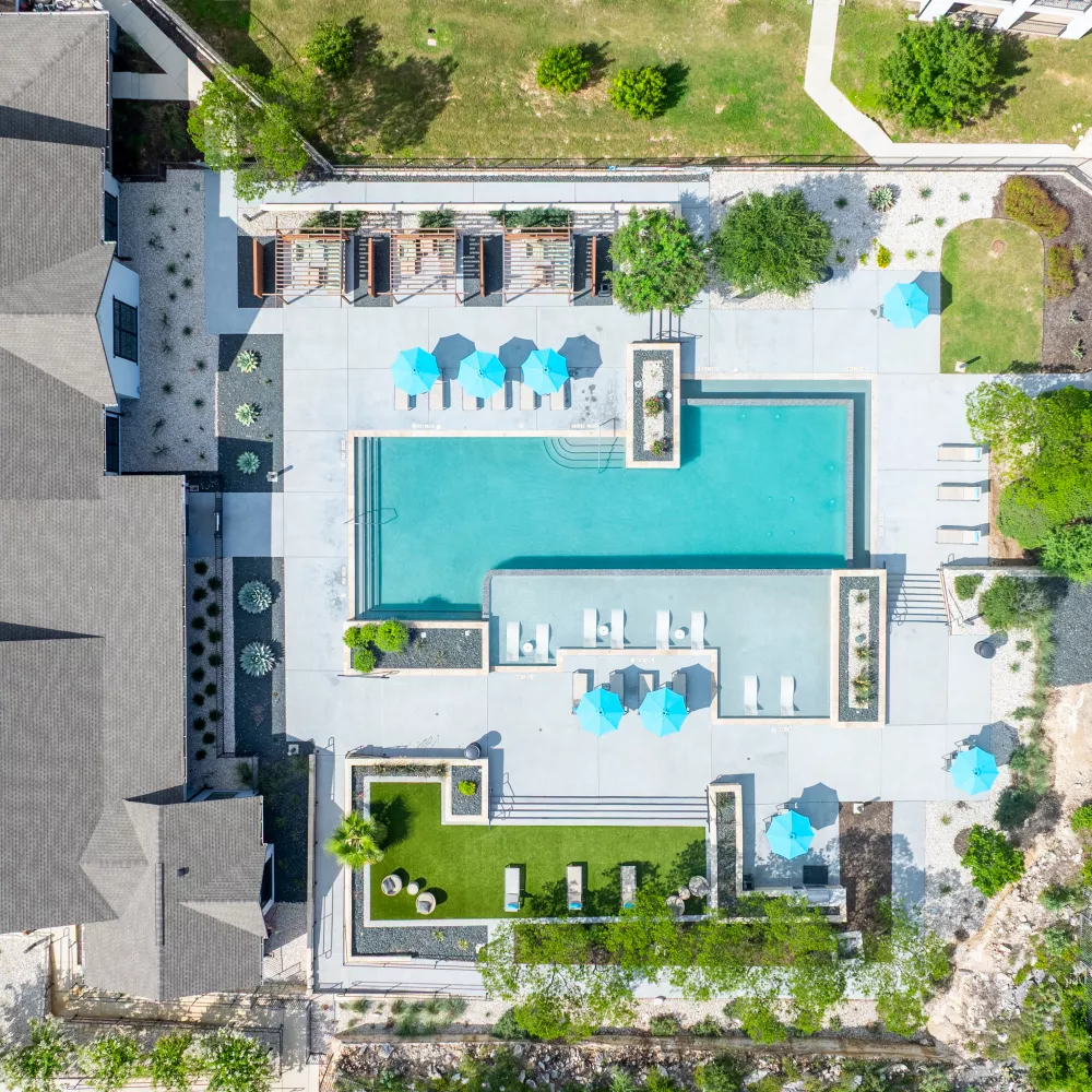 Aerial photograph: Outdoor swimming pool with blue umbrellas, sun loungers, and green lawns, next to a modern building.