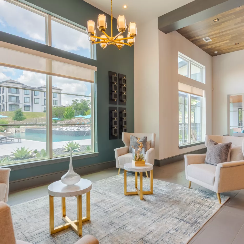 Bright, modern lobby with light armchairs, gold tables, and a chandelier, overlooking a pool and buildings.