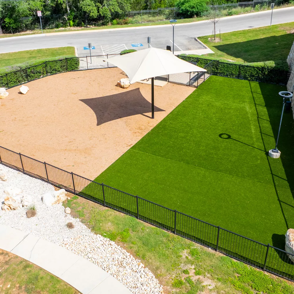 Aerial view of a dog park with sand and artificial turf sections, shaded by canopies and bordered by a stone wall.