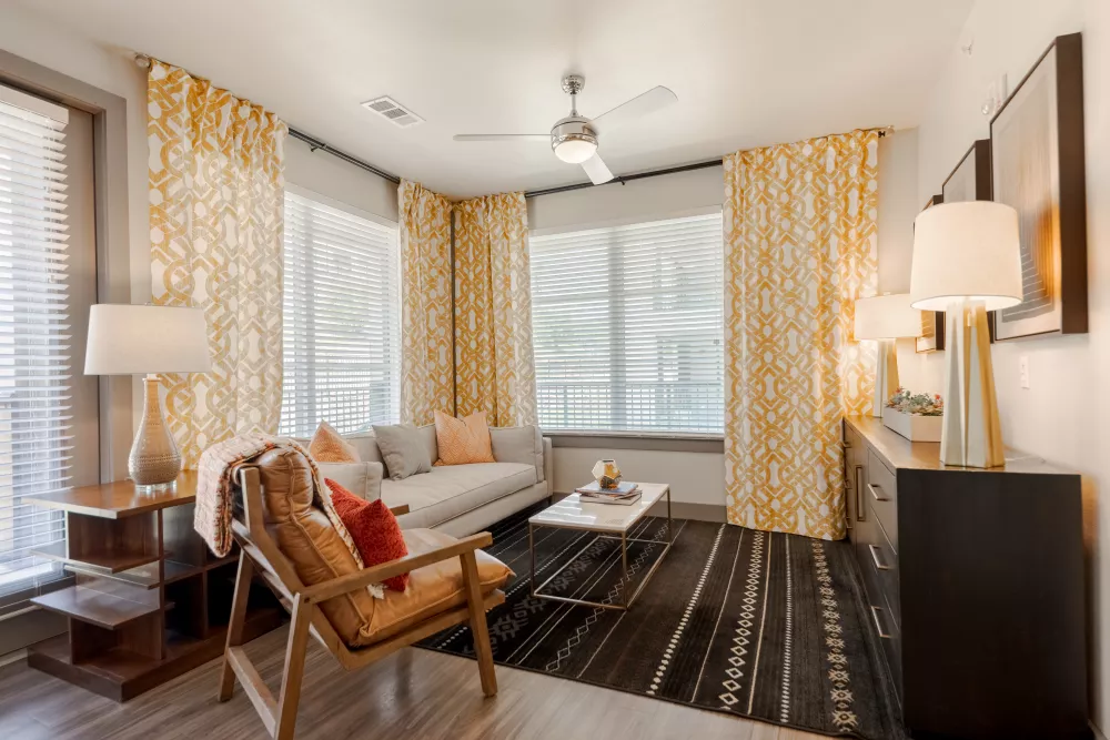 Bright living room with a white sofa, a wooden chair, a dark patterned rug, and yellow and white patterned curtains.