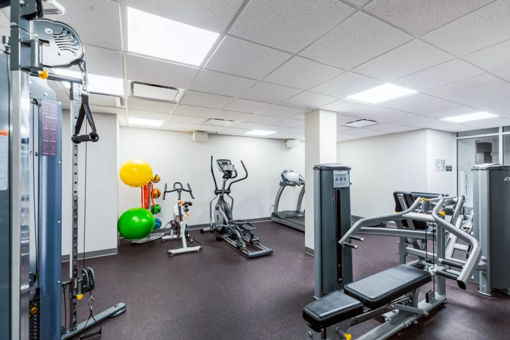 Well-lit modern gym with a cable machine, elliptical, treadmill, weight bench, stability balls, and free weights.