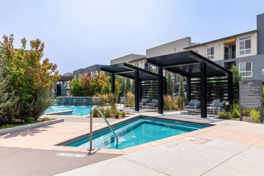Modern apartment complex pool area with hot tub, pergolas, lounge chairs, and lush landscaping.