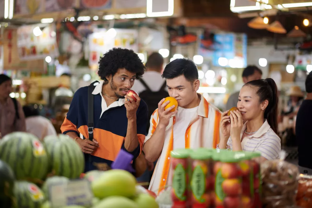 Three people, two men and one woman, smell apples and oranges at a vibrant market. Fresh produce fills the foreground.