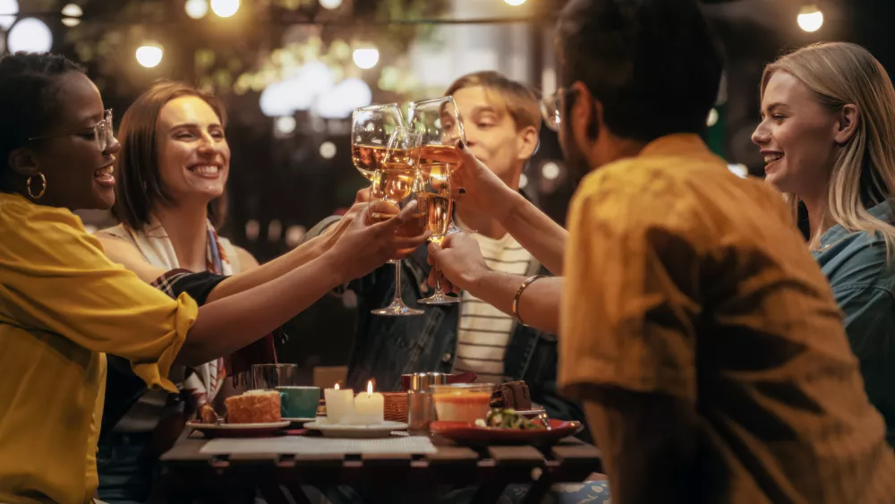 Diverse friends smiling and toasting drinks at an outdoor restaurant under warm string lights.