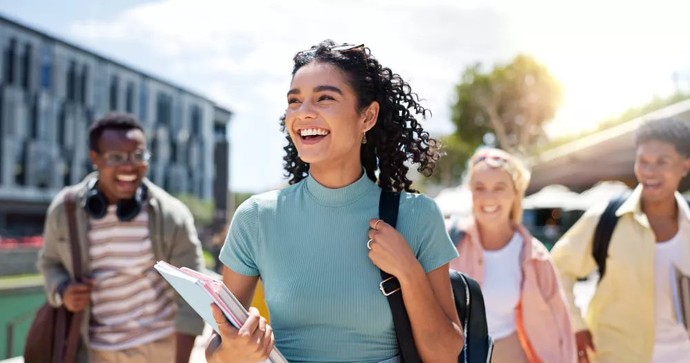 Joyful diverse college students walk outdoors on a sunny campus. A smiling woman in the foreground holds books.