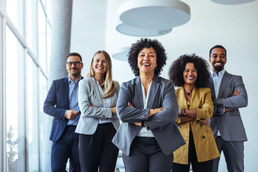 Five diverse business people smiling confidently, arms crossed, in a modern office.