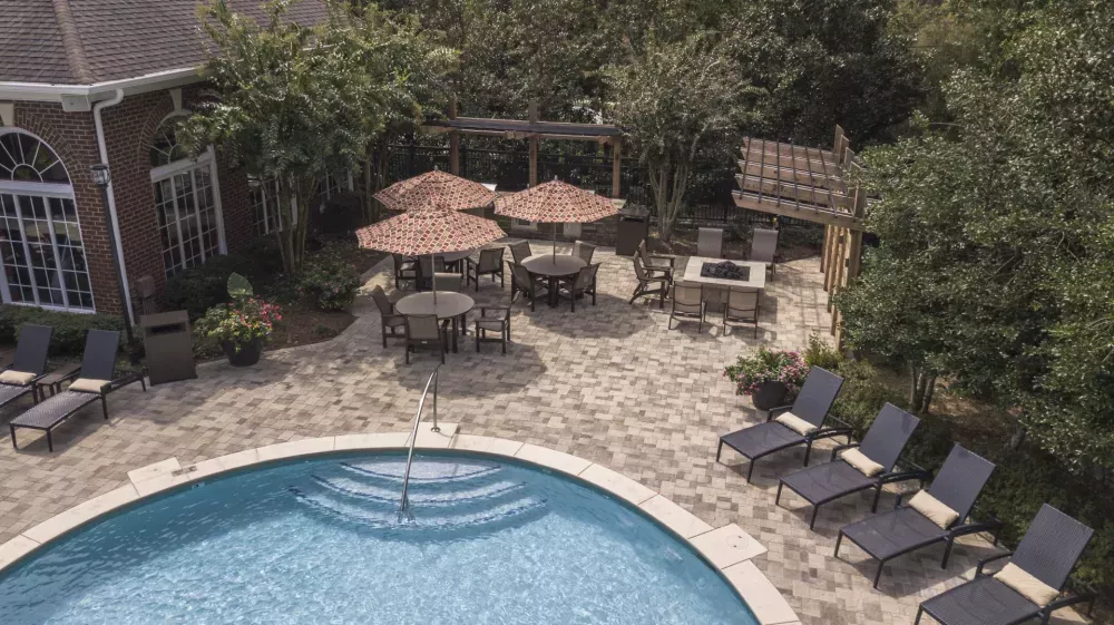 Aerial view of a resort-style pool with lounge chairs, a stone patio with shaded tables, a fire pit, and a brick building.