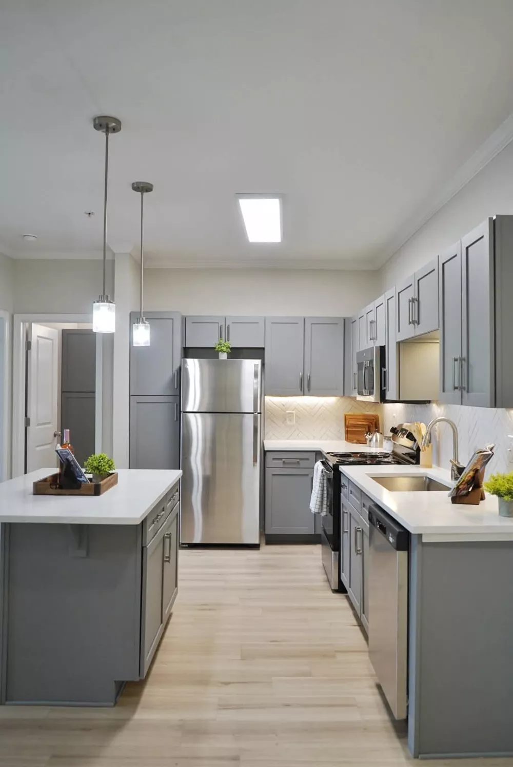 A bright, modern kitchen features gray cabinets, stainless steel appliances, white countertops, light wood floors, and an island.