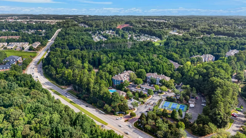 Aerial view of a highway next to apartment buildings with a pool and tennis courts, surrounded by dense green forest under a blue sky.