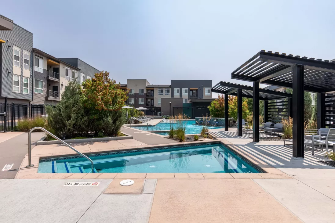 Modern apartment complex outdoor area featuring a swimming pool, hot tub, pergolas, lounge chairs, and buildings under a clear sky.