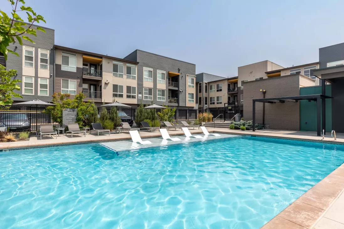 Sunny outdoor swimming pool with in-pool loungers, surrounded by modern apartment buildings with balconies.